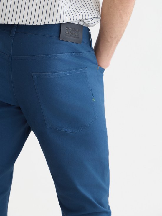 Detail van de achterkant van een blauwe chino broek met merklabel en zak.