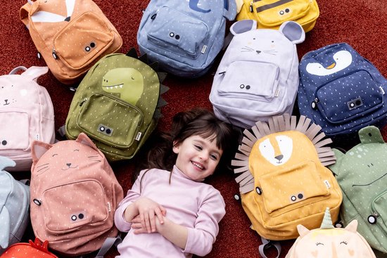 Een kind ligt tussen een verzameling kleurrijke kinderrugzakken met dierendesigns: leeuw, olifant, muis, eend, vos, dinosaurus, kat, konijn, nijlpaard, eenhoorn.