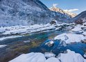 Een besneeuwd landschap met een rivier en bomen, bergtoppen op de achtergrond.