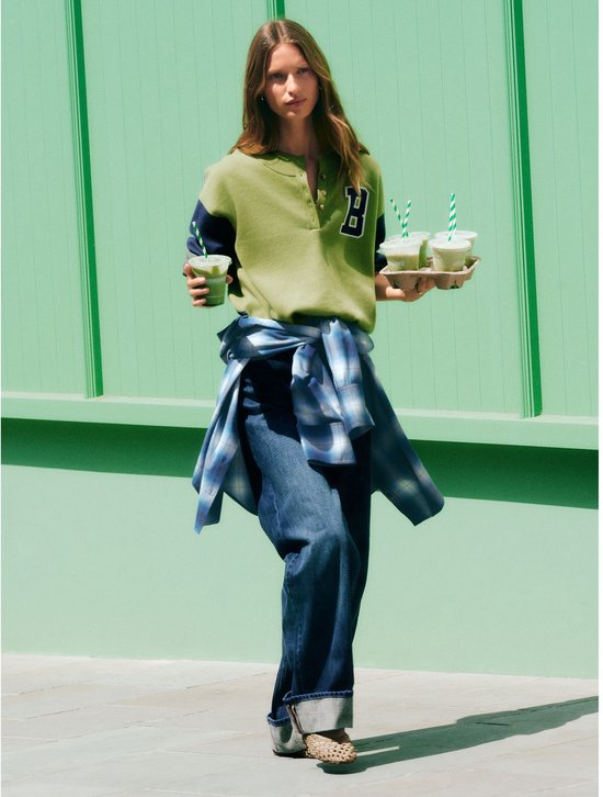 Model in een donkerblauwe boyfriend jeans met omgeslagen pijpen, een groen shirt en draagt een dienblad met drankjes.