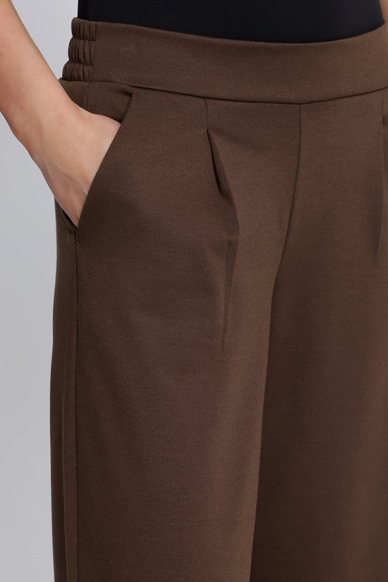 Close-up van een bruine wijde broek met plooien bij de taille en een steekzak.