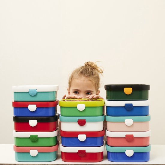 Une fille avec un chignon regarde par-dessus une pile de boîtes à lunch Mepal colorées.