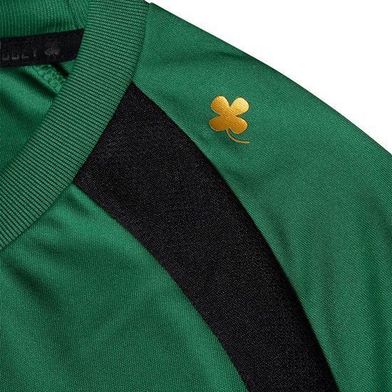 Detail van de rechterschouder van een groen sportshirt met zwarte accenten en een gouden klaver-applicatie.