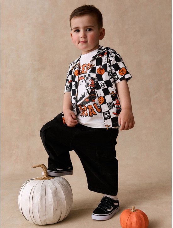 Jongen draagt een Halloween-set met ruitjeshemd vol spoken en pompoenen, en een zwarte broek, staand bij een pompoen.