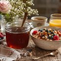 Glazen pot met donkere honing, naast een kom muesli met fruit en noten, op een houten tafel.
