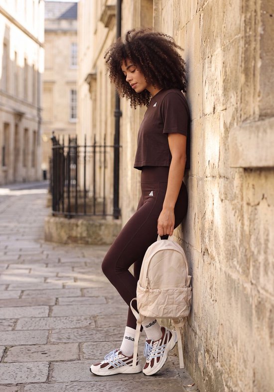 Model met bruine Adidas legging en crop top, leunend tegen een stenen muur, met beige rugzak.