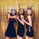 Drie vrouwen met glittermaskers en kronen, dansend met cocktails.