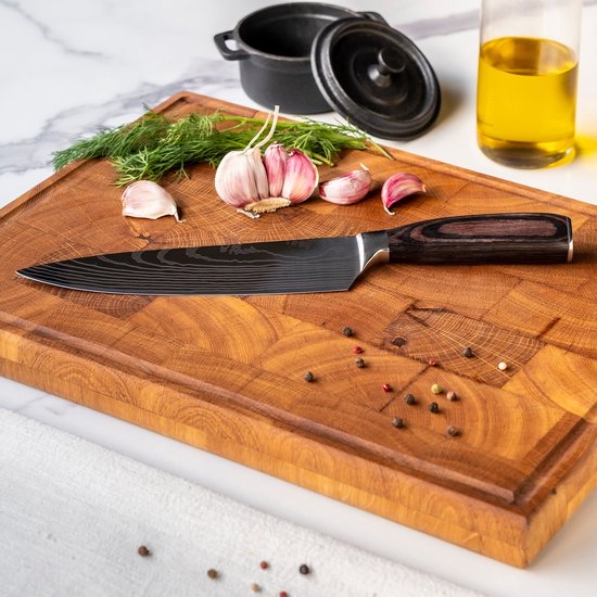 Couteau de chef avec manche marbré brun foncé et lame noire à motif damassé, posé sur une planche à découper en bois avec de l'ail et de l'aneth.