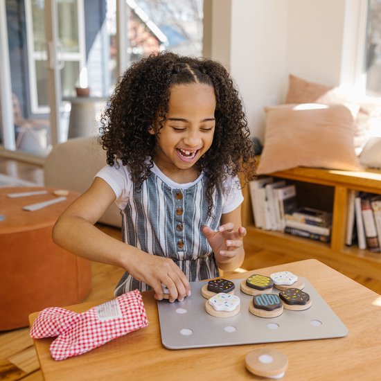 Een jong meisje met krullend haar speelt met houten koekjes op een bakplaat. Ze draagt een gestreept jurkje.