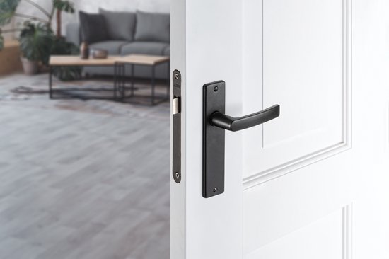 Een zwarte deurklink met schild op een witte deur, in een woonkamer.
