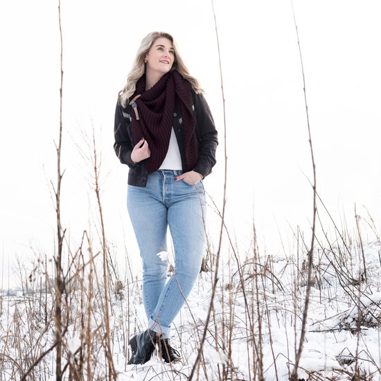 Vrouw in een winterse setting met een bordeauxrode gebreide driehoekssjaal om haar nek, gedragen met een zwarte jas en jeans.
