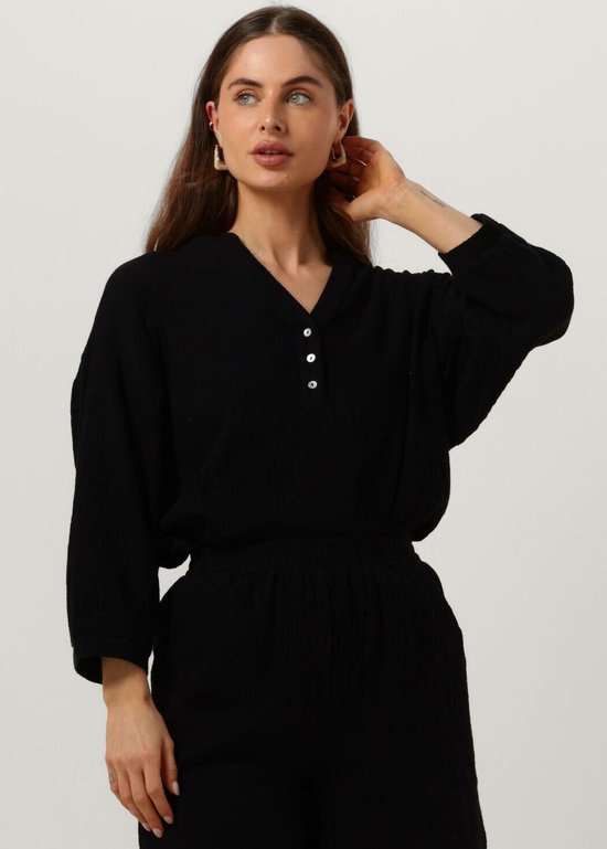 Een model draagt een zwarte, lange mouwen blouse met een korte knoopsluiting. Ze raakt haar haar aan.