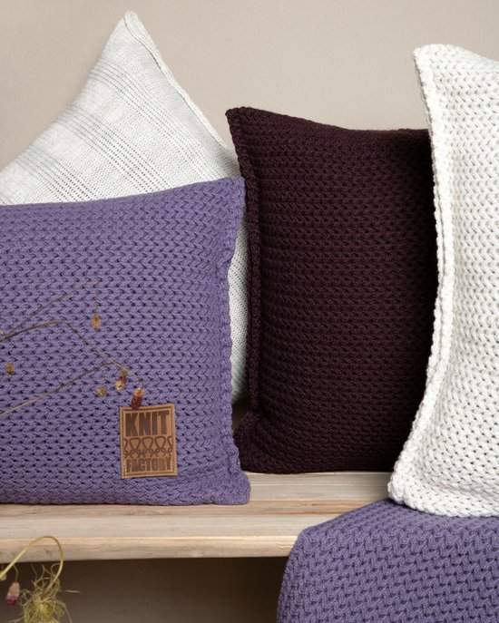 Coussins tricotés violet, bordeaux et blanc avec des étiquettes marron, partiellement sur un banc en bois.