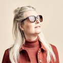 Vrouw met grijs haar, draagt een rode coltrui en een bruine zonnebril met schildpadmontuur.