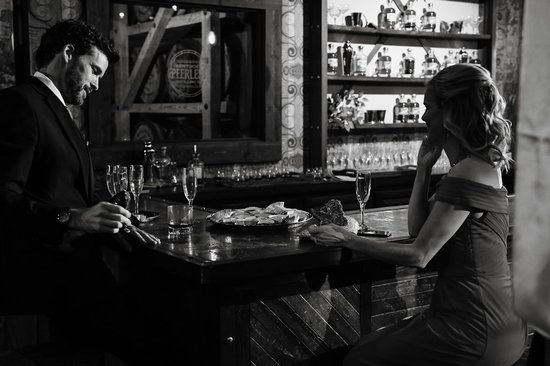 Man in pak en vrouw in rode jurk aan een bar, met drankjes en hapjes op de toonbank.