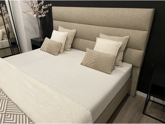Beige boxspring met gestoffeerd hoofdbord, luxe kussens en beige deken met geometrisch patroon.