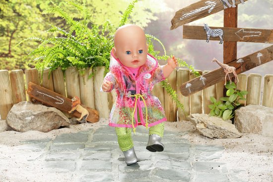 Baby pop draagt roze 'BE WILD' shirt, groene broek, zilveren laarsjes en een doorzichtige regenjas met regenboogprint.