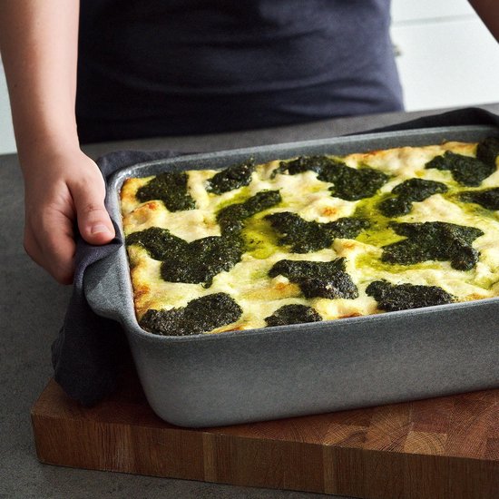 Grijze ovenschaal met lasagne, wordt met een donkergrijs linnen doek door iemand vastgehouden.