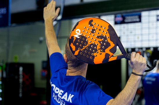 Persoon die een padelracket omhoog houdt, met een oranje en zwart camouflagepatroon en een zwart handvat.