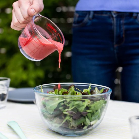 Een glazen maatbeker met rode dressing die wordt uitgegoten in een glazen kom met gemengde salade.