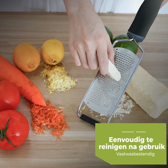 Râpe en acier inoxydable avec manche noir utilisée pour râper un morceau de fromage, avec carotte, citron, tomate sur une planche en bois. Lave-vaisselle.