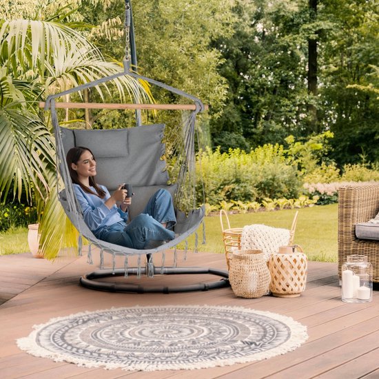 Grijze hangstoel met franjes en drie kussens, in de tuin op een houten terras.
