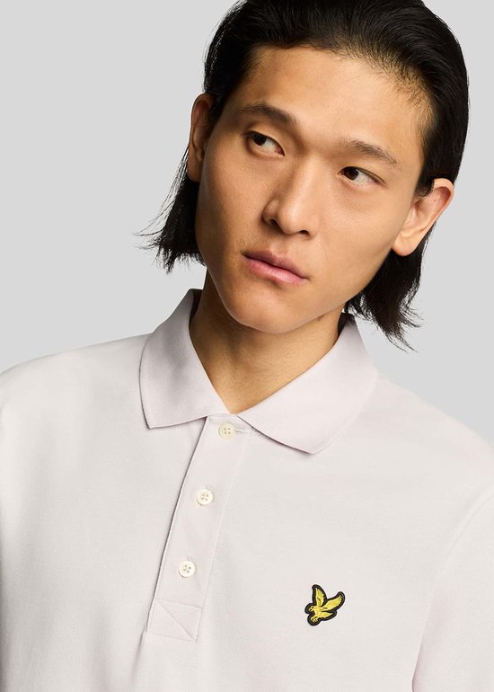 Een man met donker haar draagt een lichtpaarse poloshirt met een geel Lyle & Scott-vogellogo op de borst.