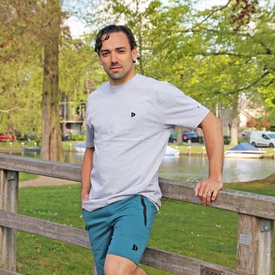 Man in lichtgrijze T-shirt met zwart logo, turquoise korte broek met logo, staand bij een houten reling.