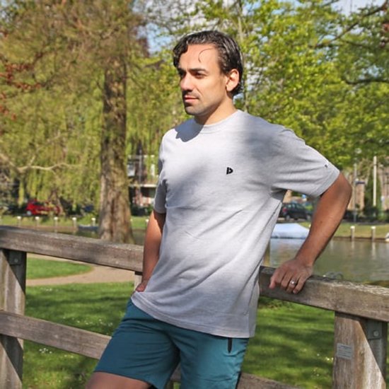 Man in lichtgrijs T-shirt en blauwgroene short, leunend op een houten reling bij een kanaal.