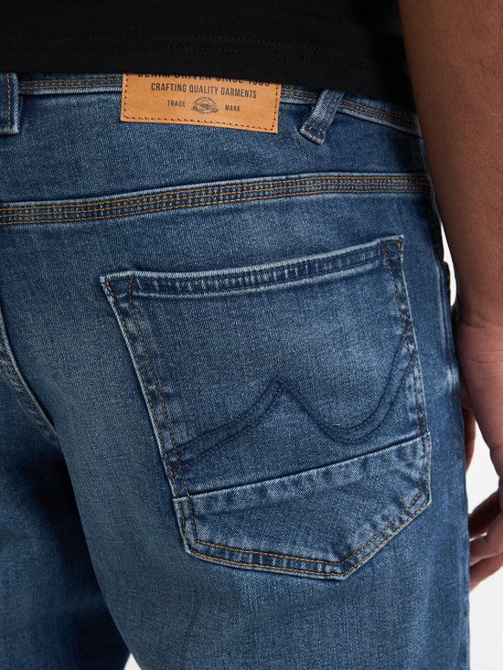 Detail van de achterkant van een blauwe jeans met opgestikte zakken en een leren label met 'Crafting Quality Garments'.