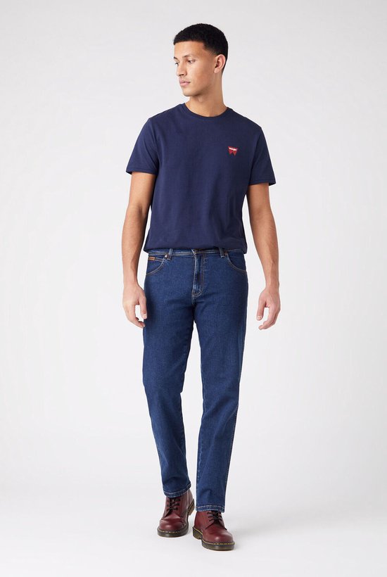 Blauwe Wrangler jeans, regular fit, gedragen met donkerblauw T-shirt met klein rood logo en rode schoenen.