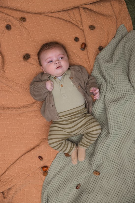 Baby in een lichtgroen broekje met streepjes en een bruin vestje, liggend op een oranje en groen plaid.