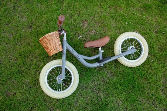 Lichtgrijze loopfiets met bruin zadel, bruine handvatten en rotan mandje, liggend op gras.