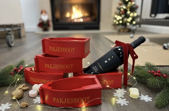 Stapel rode houten pakjesboten met de tekst "PAKJESBOOT", een fles wijn en kerstdecoratie.