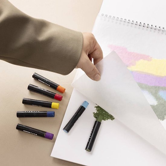 Close-up: hand tilt een tussenvel op van een oliepastelblok. Diverse oliepastels liggen op een beige ondergrond.