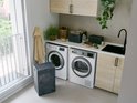 Antracietkleurige wasmand naast een wasmachine in een lichte wasruimte. De wasmand heeft een deksel en een opengewerkt patroon.