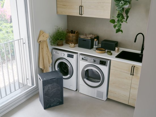 Antracietkleurige wasmand naast een wasmachine in een lichte wasruimte. De wasmand heeft een deksel en een opengewerkt patroon.