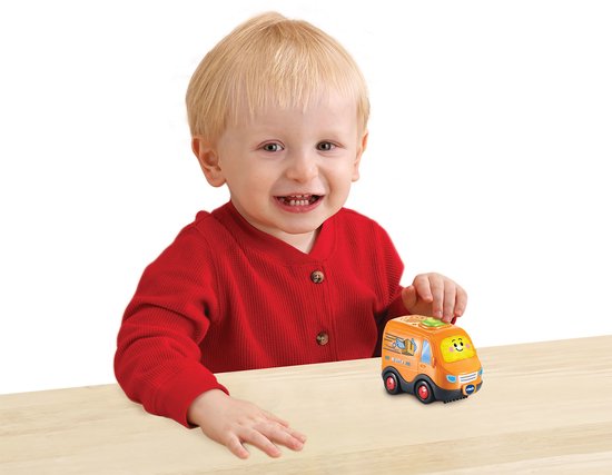 Jongetje in rode trui kijkt lachend naar een oranje VTech Boris Bestelbus speelgoed auto op een houten tafel.