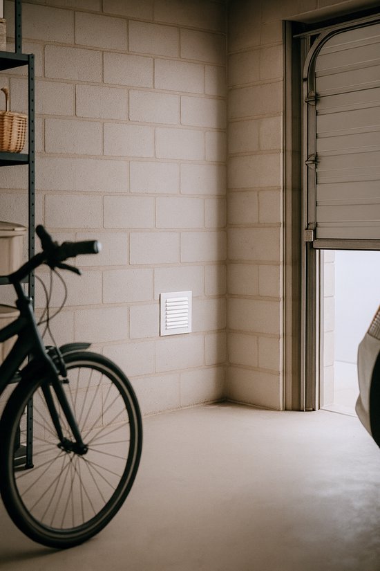 Wit ventilatierooster met lamellen op een betonnen muur, naast een fiets.