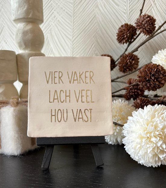 Crèmekleurige spreuktegel met gouden tekst: 'Vier vaker, lach veel, hou vast'. Staat op een zwart schildersezel.