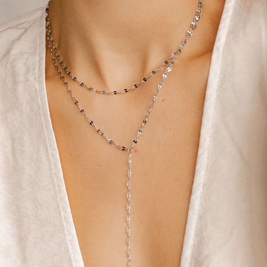 Close-up van een vrouw die een gelaagde zilverkleurige ketting met fijne schakels en een Y-vormige drop draagt.