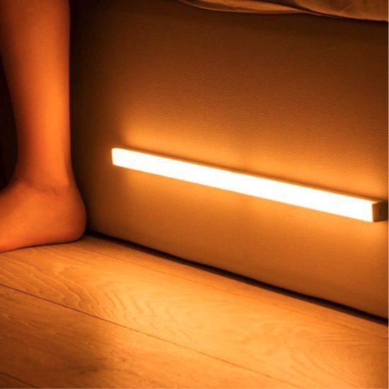 Een enkel been naast een oplaadbare, magnetisch te monteren LED-strip die warm wit licht uitstraalt op een houten vloer.