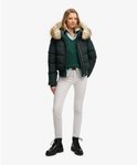 Vrouw in donkergroene bomberjack met bontkraag, groen kabelvest en witte jeans.