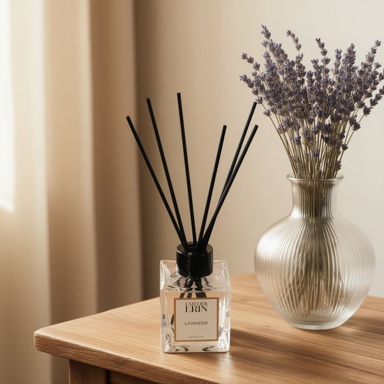 Een glazen geurverspreider met zwart riet, met het label 'L'ATELIER ERIN LAVENDER'. Naast een vaas met gedroogde lavendel op een houten tafel.