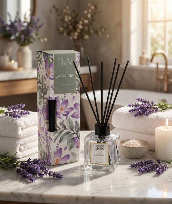 Een glazen geurverspreider met zwart riet, met het label 'L'ATELIER ERIN LAVENDER', naast een doos met lavendelprint. In een badkamer met handdoeken, lavendel en zout.