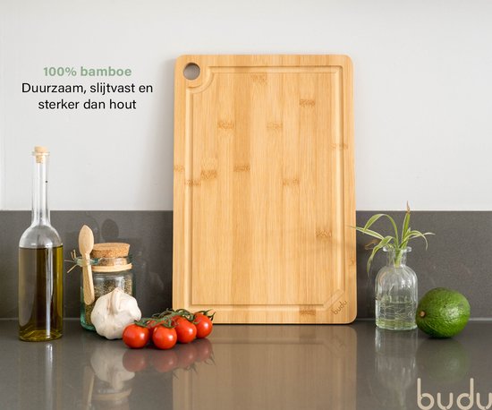 Een Budu bamboe snijplank op een aanrecht, met een fles olijfolie, knoflook, tomaten en een plantje ernaast.