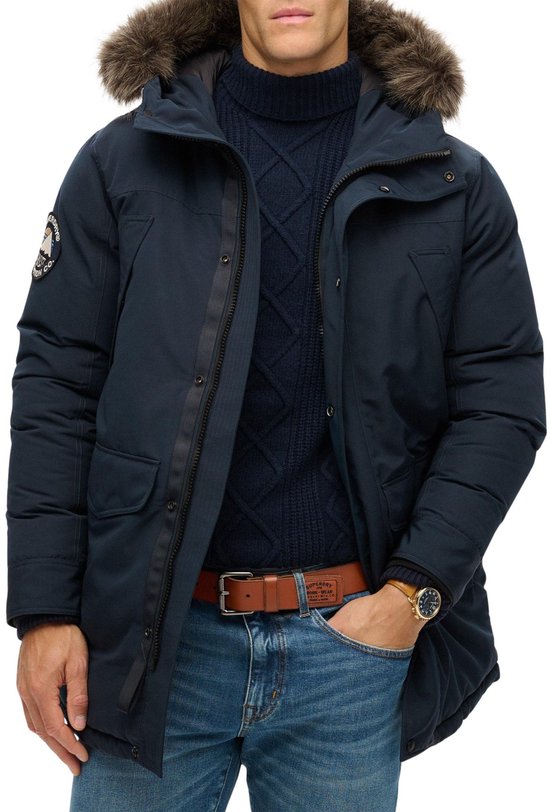 Donkerblauwe parka met bontkraag, gedragen door een man met spijkerbroek, riem en horloge.