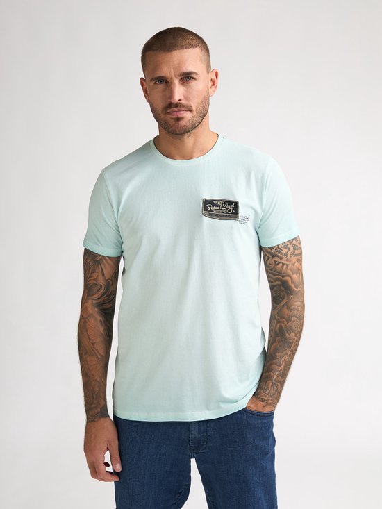 Man met kort groen T-shirt met logo-patch op de borst en spijkerbroek.