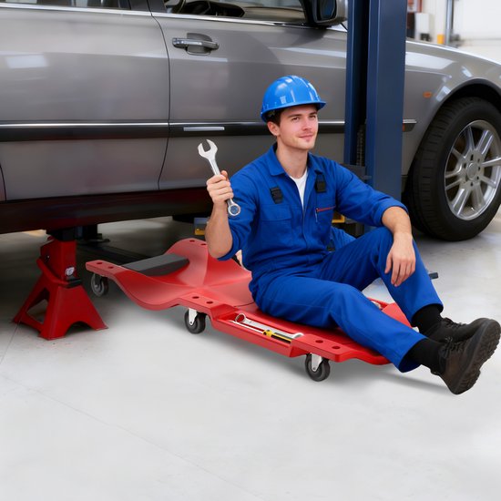Monteur in blauw uniform, helm op, houdt moersleutel vast terwijl hij op rode rolplank zit onder auto.