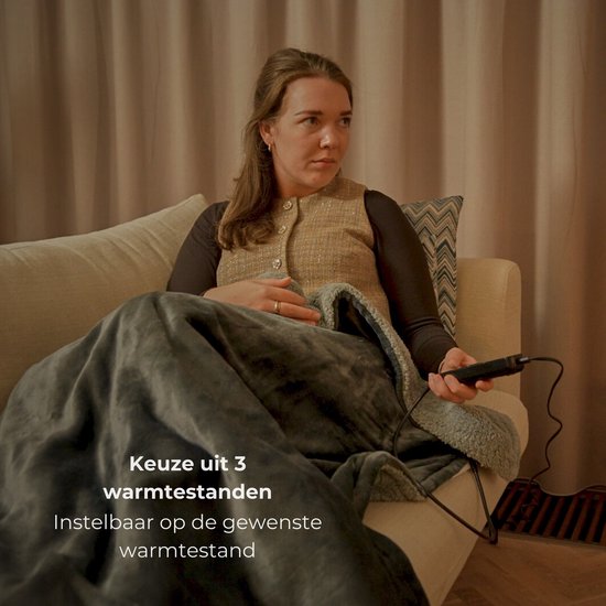 Vrouw op een bank, bedekt met een grijze elektrische deken, die de afstandsbediening vasthoudt.